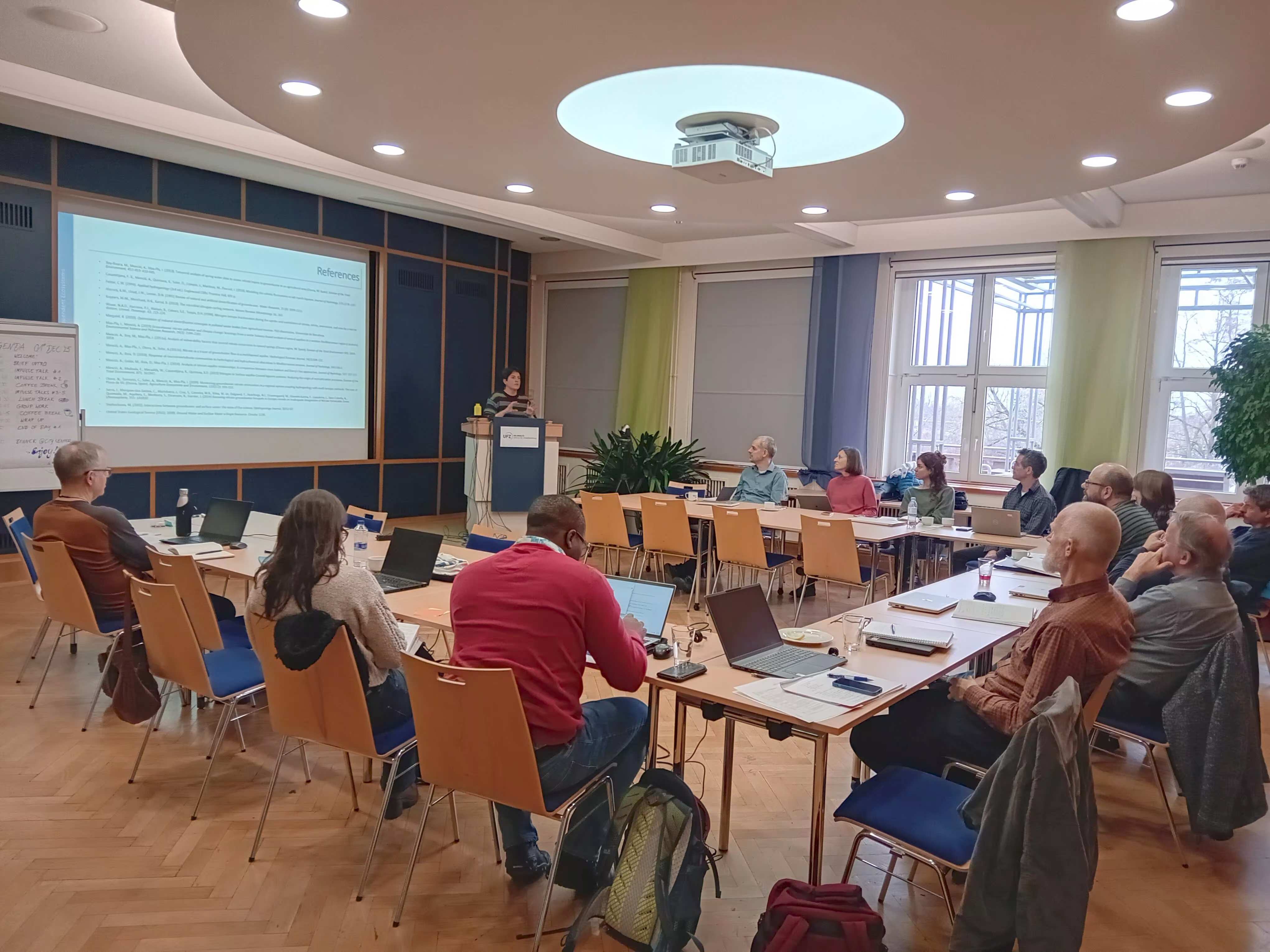 13 scientists are sitting at tables with notebooks. They are looking to the front of the room where a scientist ist helding a presentation.