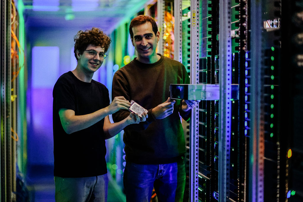 Zwei m&auml;nnliche Personen pr&uuml;fen Bauteile in einem Serverraum und l&auml;cheln in die Kamera. Foto: Anne Schwerin